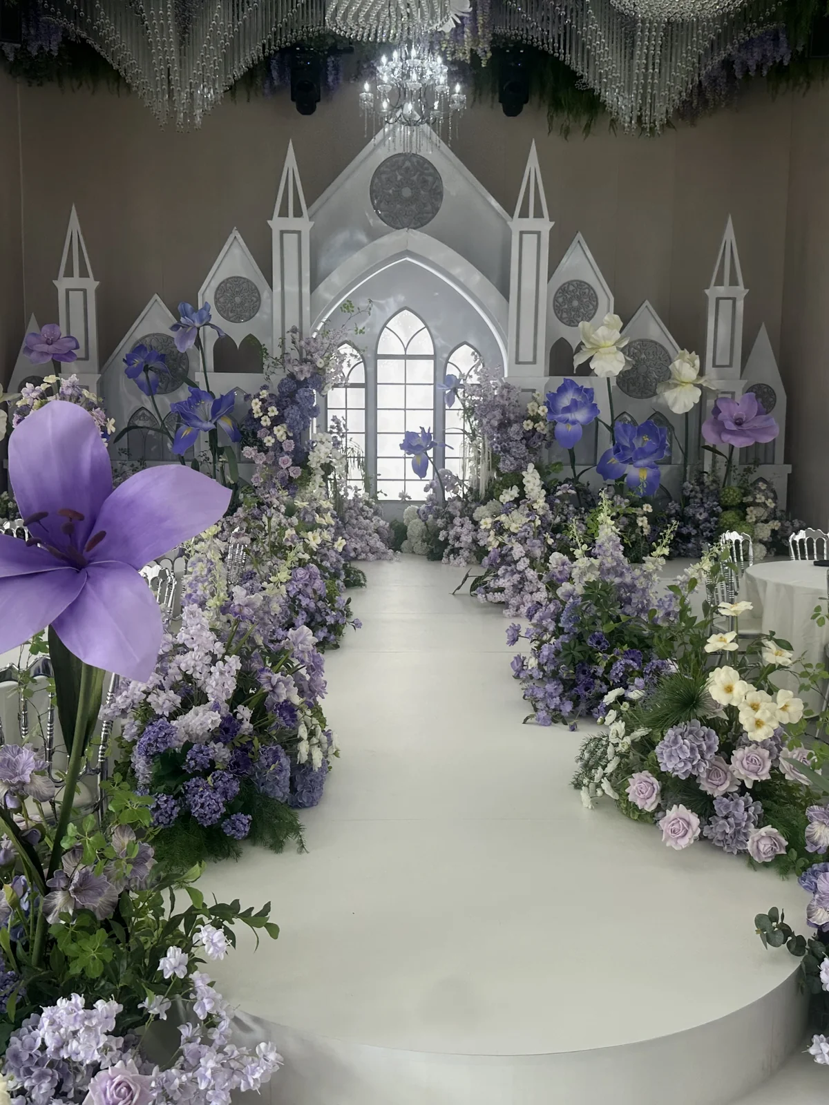Purple and white artificial flowers lining a wedding ceremony aisle with a church-style backdrop