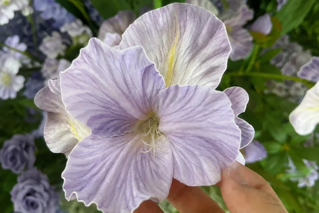 A hand holding a pale purple artificial iris with visible petal veining and a yellow center.