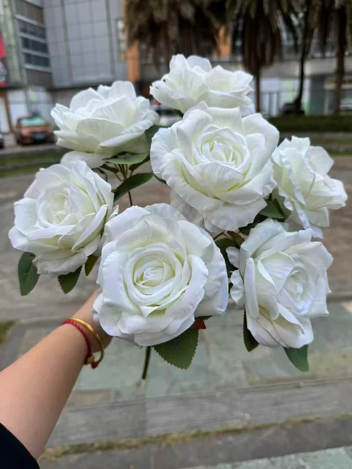 A hand holding a bunch of white artificial roses outdoors, showing layered petals and green leaves.
