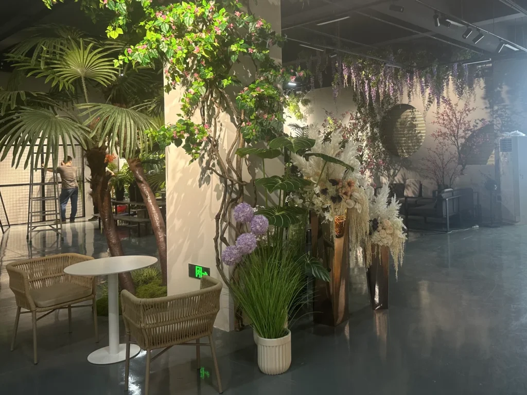 A table and wicker chairs among artificial greenery and floral displays in the PJTL Foshan showroom.