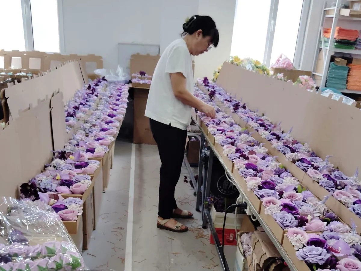 A worker handling rows of purple and lilac artificial flower arrangements in open cardboard boxes.