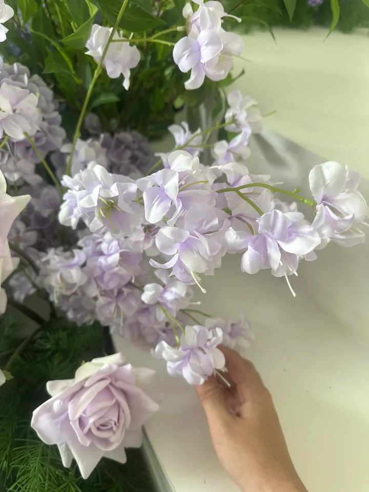 A hand holding a pale lilac artificial flower spray, showing fabric petals and green stems.