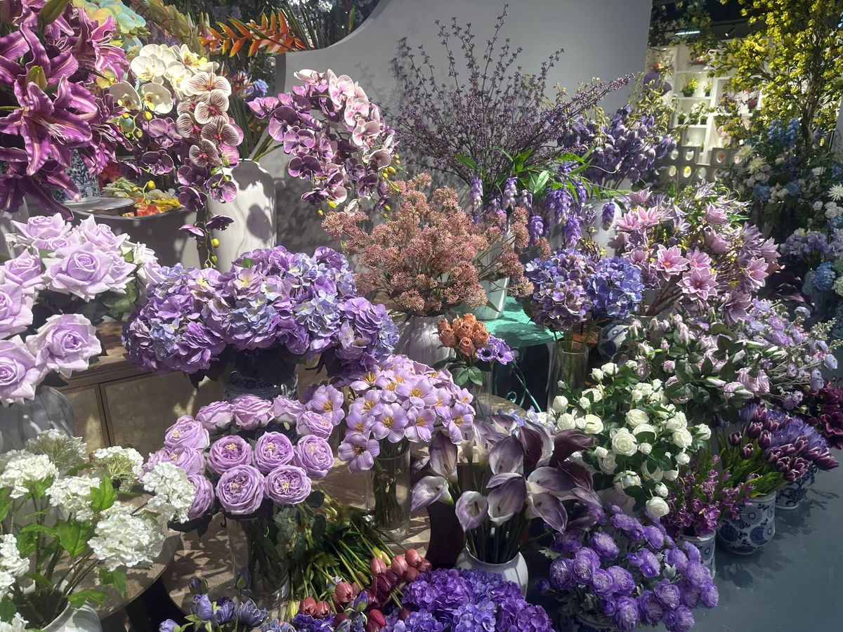 Purple and lilac artificial roses, hydrangeas and orchids arranged together in showroom vases.