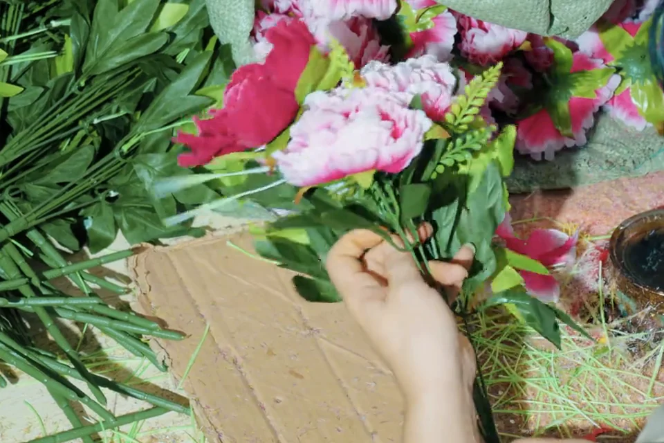 A worker assembling pink artificial flowers and green stems at a worktable.