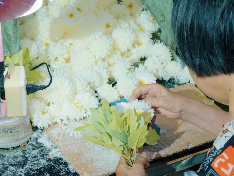 Hands assembling white artificial flowers with pale green leaves beside loose flower components.