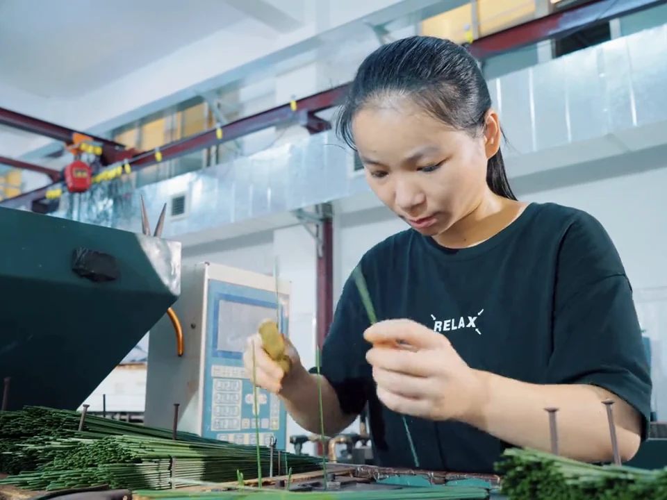 A worker positioning green artificial flower stems at a production workstation.